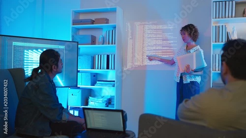 A professional East Asian female software engineer stands holding a laptop and gesturing toward lines of code projected on a wall display, presenting data scripts to her multiethnic colleagues during 