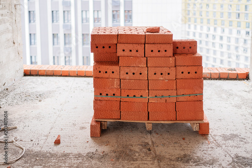 Foto A stack of red perforated clay bricks prepared on a makeshift pallet on a concrete floor inside an unfinished high-rise building with an urban cityscape view