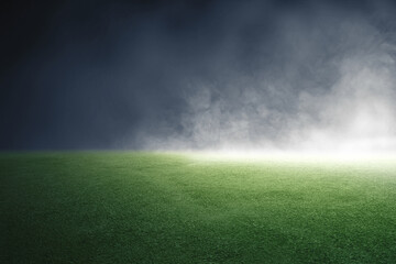 An empty green grass turf field illuminated by a bright spotlight under a dark, smoky, and misty night atmosphere, ideal for sports or event background