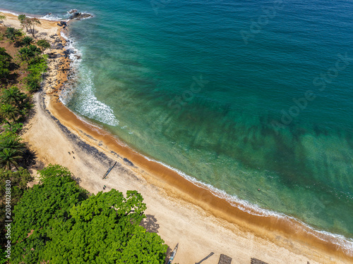Aerial view of a tropical coastline with turquoise ocean waves washing onto a sandy beach lined with lush green trees in Monrovia, Liberia.