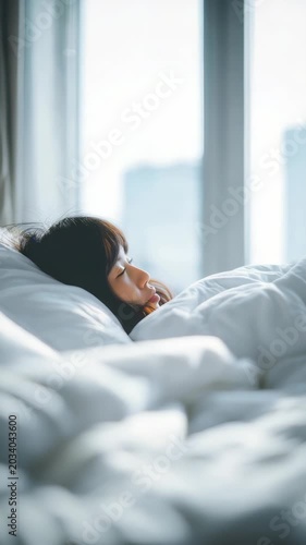 A peaceful morning scene shows a woman relaxing in bed, surrounded by fluffy white bedding and illuminated by gentle daylight. She appears calm and reflective, enjoying a quiet moment indoors