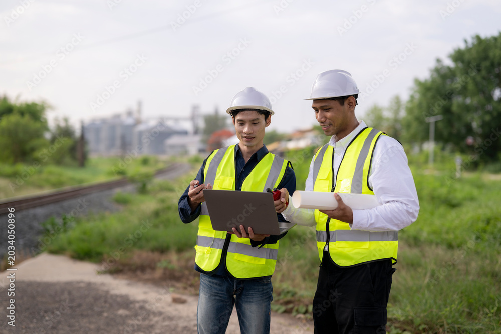 Obraz premium Two construction engineers in safety gear using tablet and clipboard outdoors near railway tracks