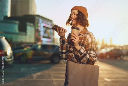 Quadro su tela Woman holding coffee and phone with a bright smile on an urban street at golden hour sunlight, beanie and tote in a candid scene of authenticity, mindful living and emotional storytelling