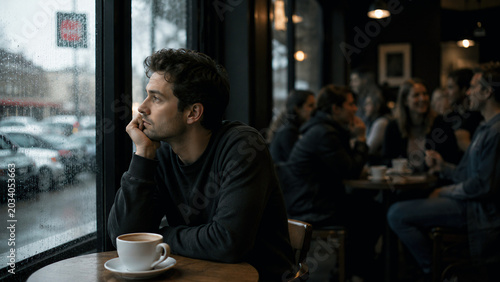 Lonely Young Man Looking Out Rainy Window in Busy Coffee Shop