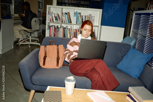 Young adult Caucasian woman studying on laptop in library lounge, researching coursework and online learning. Useful for education marketing, student life, academic services
