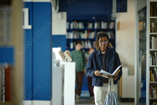 Young Black male student reading book while walking through library aisle, studying independently for academic success
