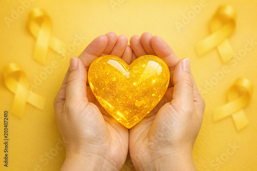 A human hand gently cradling a heart-shaped object filled with yellow glitter, surrounded by small yellow ribbons. A symbol of love and support in the context of health or wellness awareness.