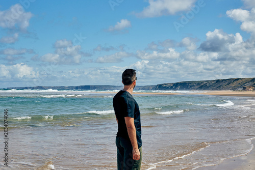 Φωτογραφία Young man standing on a sandy beach looking at the scenic ocean coastline and wa