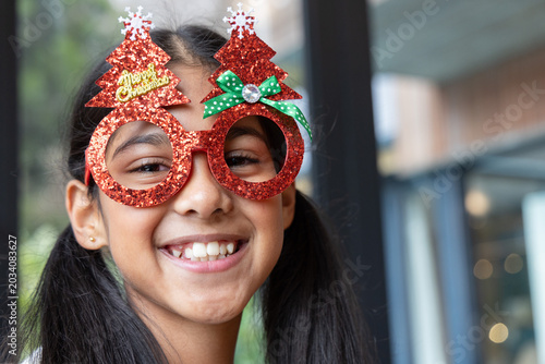 Indian female child smiling and wearing red glitter Christmas-tree glasses at cafe windows
