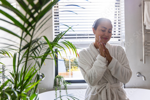 Asian woman doing skincare in bathroom wearing robe by freestanding tub with plants, copy space