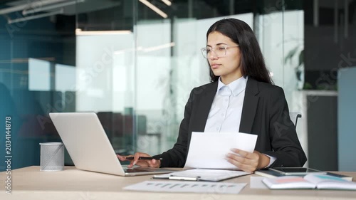 Thoughtful young businesswoman reviewing financial reports while working on laptop at desk in business office. Female accountant is focused on analyzing data, preparing documents, tax calculations.