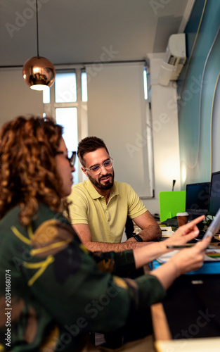 Man with glasses sitting at an office desk, smiling and passing documents to a female colleague during a work discussion.
