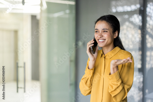 Billede på lærred Smiling businesswoman talking on a mobile phone in a modern corporate hallway, c