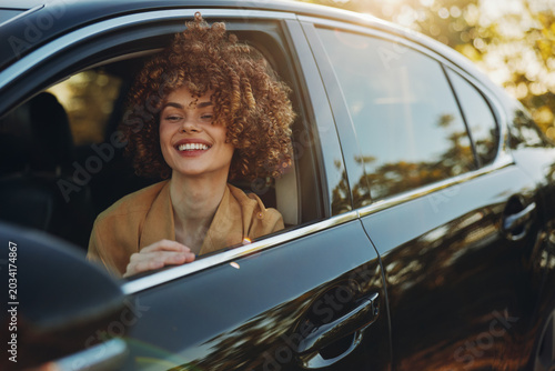 Tableau sur toile A smiling woman leans from a car window during golden hour, embracing a carefree street style
