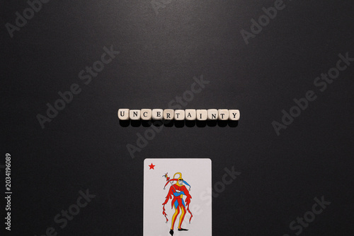 Tableau sur toile Uncertainty spelled with letter tiles above a joker card on dark background, symbolizing unpredictability, risk, instability and doubt