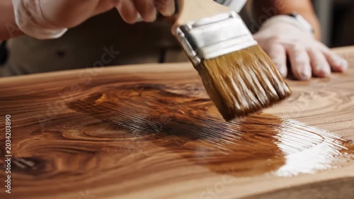 Close-up of hands applying wood finish with a brush. Woodworker protecting a wooden slab, enhancing natural grain. Home renovation and interior design concept