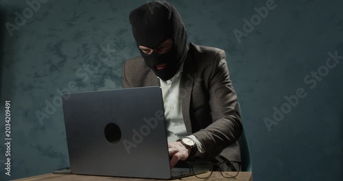 Masked man sits at laptop while typing during online scam operation. Fraudster uses computer as hiding identity behind balaclava and dark clothing in room