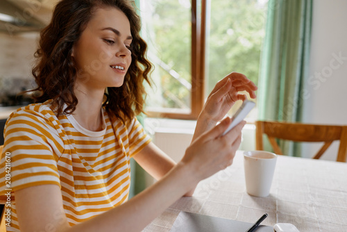 Billede på lærred Young woman with wavy brunette hair sits at a bright home table, scrolling on a