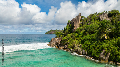 Turquoise waters meeting rocky cliffs surrounded by dense, lush greenery under a bright sky. Seychelles, Mahe.