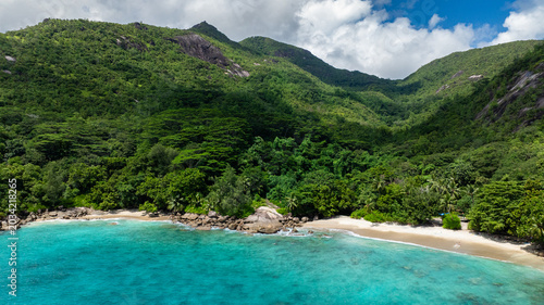 A small sandy beach with clear turquoise water surrounded by dense green vegetation and rocks. Seychelles, Mahe. Anse Major.