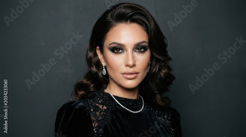 A woman with dark hair and makeup, wearing a black lace dress, standing against a dark background.