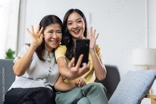 Photos Asian mother and adult daughter enjoying a virtual video call together from their home living room couch, waving hands, and smiling while staying connected with technology