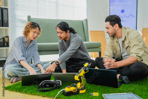Diverse team of young tech engineers collaborating on robotics development and programming a yellow robotic arm, using laptops while sitting together in a modern startup office workspace, focusing on 