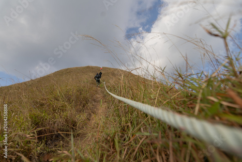 A long-distance view of a lone hiker walking along a narrow, grassy mountain ridge path, guided by a prominent safety rope in the foreground, with layers of hills fading into the background.