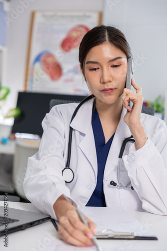 Doctor stethoscope smartphone clinic medical consultation female physician white coat using smartphone while writing notes clinic desk, focused expression during telemedicine call Vertical