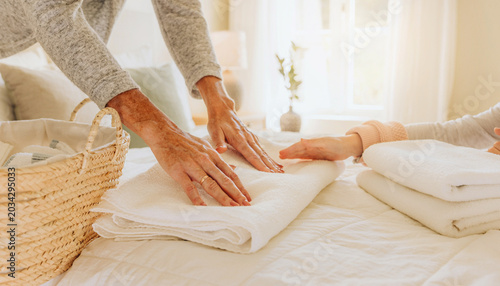 A mother and daughter’s hands work together to fold clean white laundry on a bed in a sunlit bedroom.