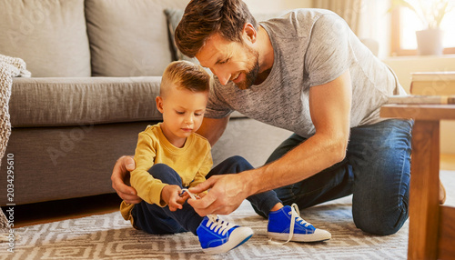 A father teaches a young child how to tie shoelaces on a pair of bright blue sneakers at home.