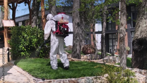 Worker in protective suit spraying garden with pesticide using backpack fogger, outdoor pest control treatment in residential yard