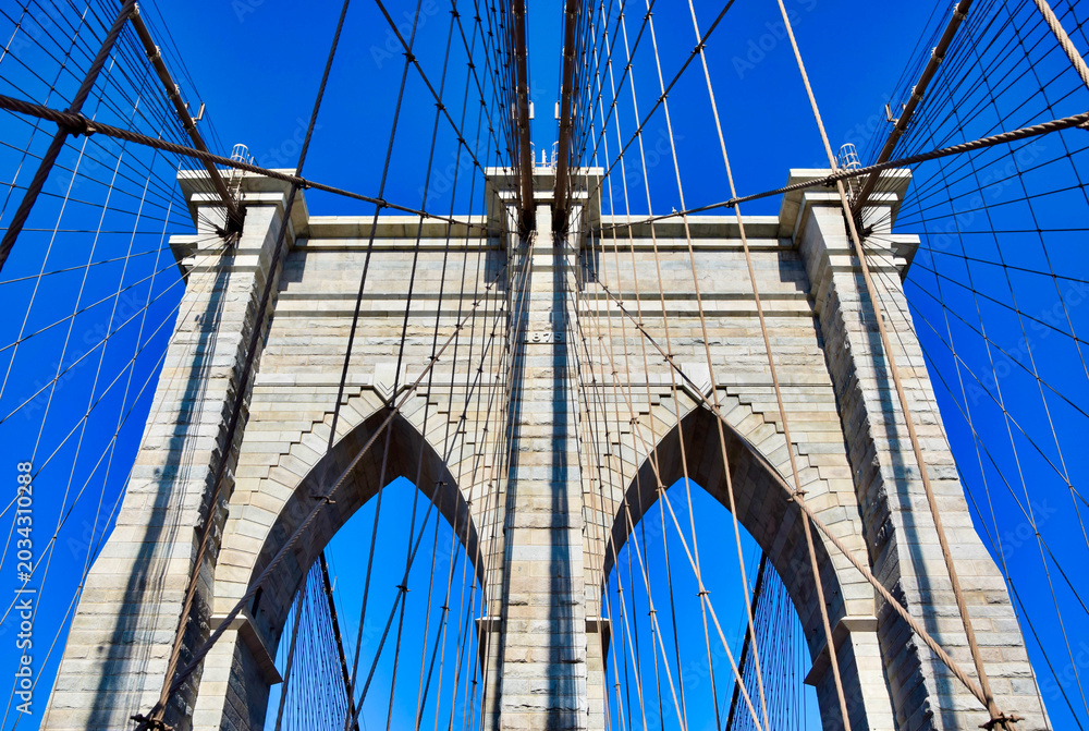 Fototapeta premium Brooklyn Bridge granite tower with gothic arches and suspension cables seen from walkway against vivid blue sky, close-up architectural detail, New York City