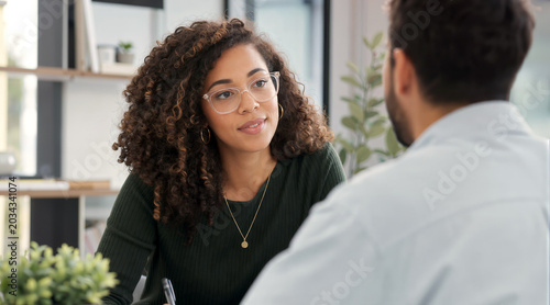A professional woman with curly hair and glasses listens intently to her colleague during a business meeting in a bright, modern office setting, showing focus and engagement.