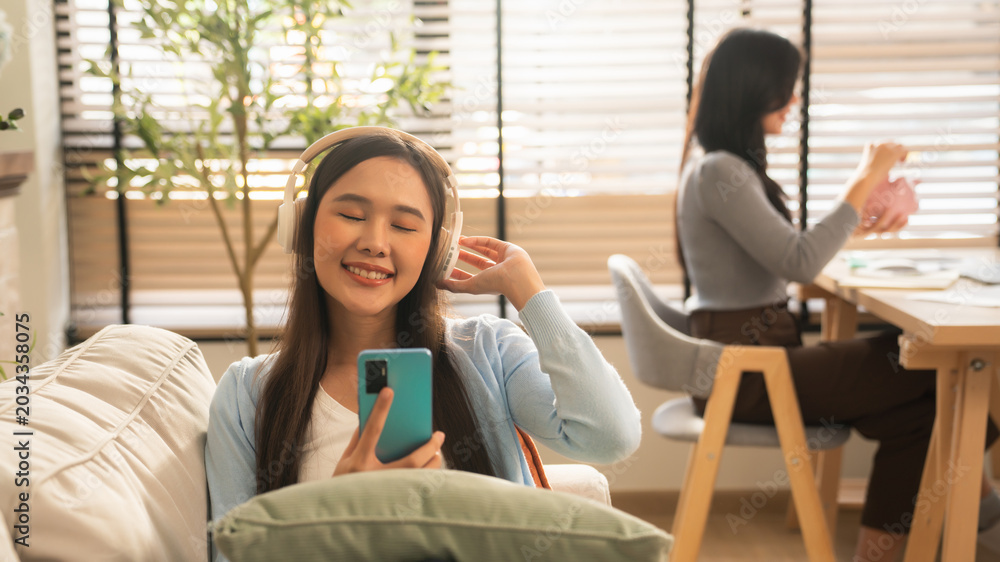 Fototapeta premium Happy Young Asian Woman Enjoying Music with Wireless Headphones and Smartphone, Smiling and Relaxing on Cozy Sofa in Modern Living Room.