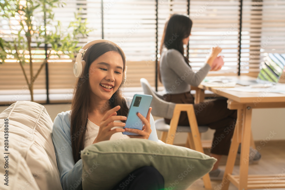 Fototapeta premium Happy Young Asian Woman Enjoying Music with Wireless Headphones and Smartphone, Smiling and Relaxing on Cozy Sofa in Modern Living Room.