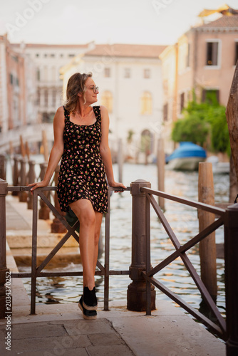 Stylish young woman in a dark floral mini dress leaning on a railing by a scenic Venetian canal at warm golden hour light in Venice, Italy