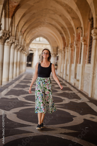 Stylish young woman in glasses walking through the elegant arched colonnade of the Doge's Palace in St. Mark's Square, Venice, Italy on a summer day