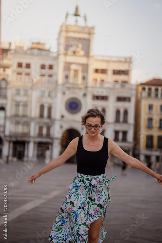 Joyful young woman in glasses spinning and dancing with arms outstretched in the historic St. Mark's Square in Venice, Italy at dusk