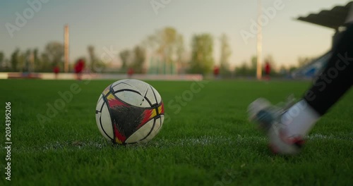 Close-up soccer ball on a green field touchline at sunset, with players training in the background and a stadium stand beside the pitch