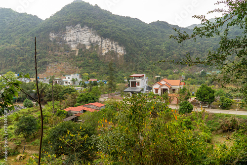 A scenic view of a rural Vietnamese village nestled at the base of lush, green mountains with a prominent cliff face