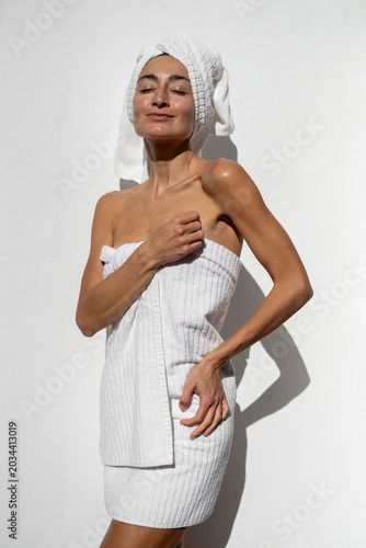 Konstfotografi Woman wrapped in white towel with hair towel standing against white wall, enjoyi