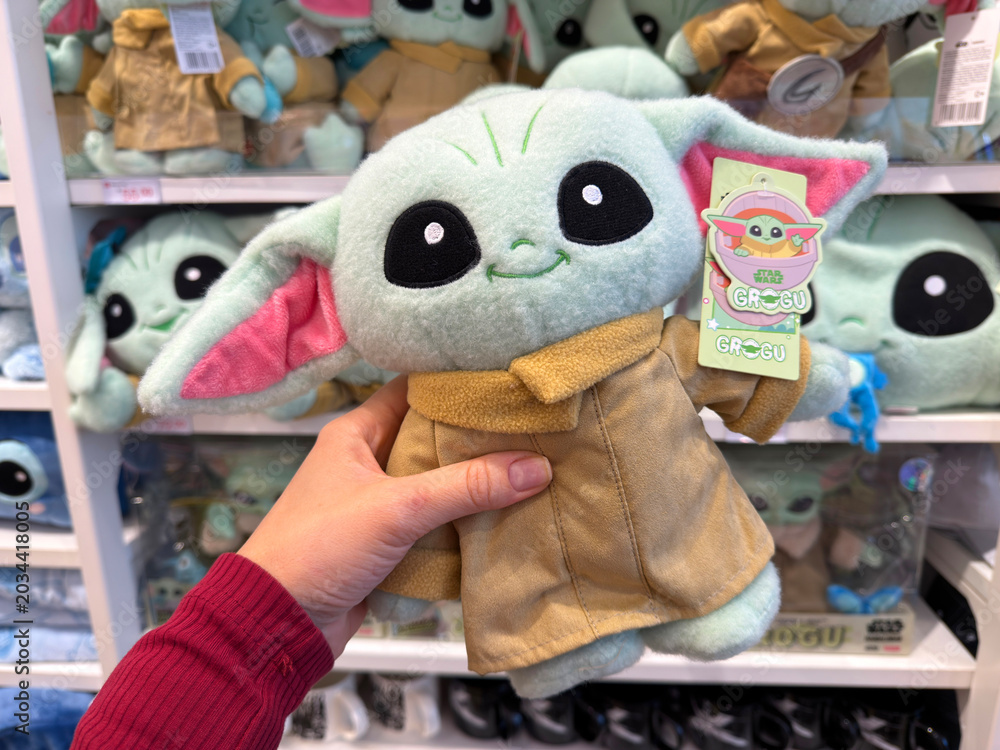 Naklejka premium Dubai, UAE - 24 may 2026: Hand holding Grogu plush character in modern toy store surrounded by collectible Star Wars merchandise, representing entertainment retail and fandom culture lifestyle