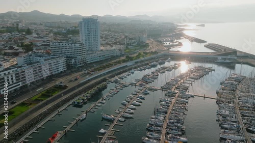 Aerial view of the modern marina in Ponta Delgada, crowded with numerous yachts and sailboats, adjacent to the bustling city coastline and waterfront promenade at sunrise, São Miguel Island, Azores.