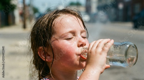 Overheated little girl without a hat drinking water from a glass bottle on a hot sunny day. Risk of sunstroke, heatstroke, and dehydration concept