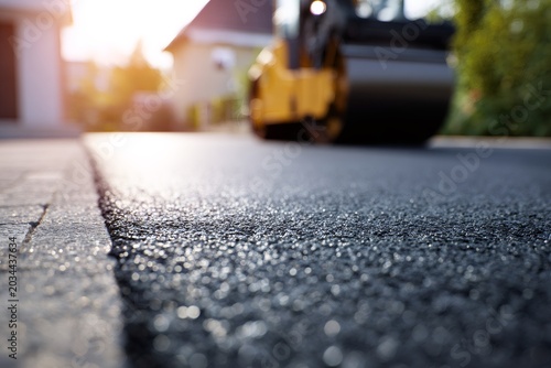 A close-up of new asphalt texture on a freshly paved road. A road roller works on a street in the background during construction