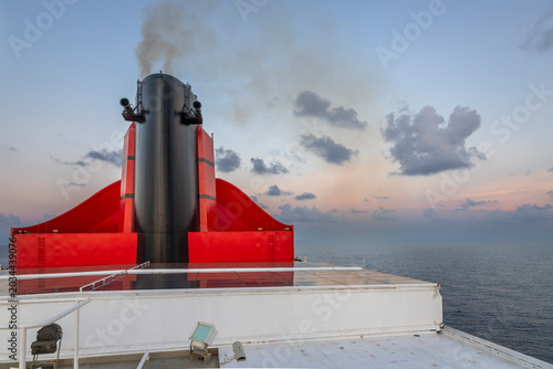 Close-up view of a large red and black cruise ship funnel releasing smoke into the sky at dusk. Captured aboard a luxury ocean liner, this maritime scene conveys the power and scale of modern cruise t