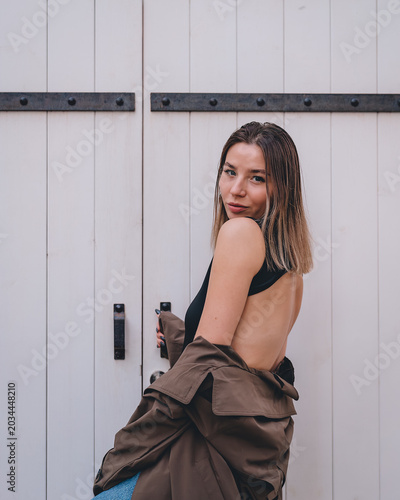 A stylish young woman stands against a backdrop of light-colored wooden doors. The woman turns, throwing a brown coat over her shoulders. A minimalist composition in a modern urban style.