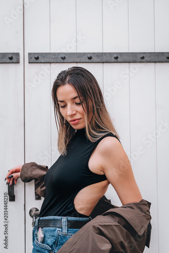 A stylish young woman poses against a white wooden door with metal hinges. She's wearing a black sleeveless bodysuit with an open back, blue jeans, and a brown trench coat draped over her shoulders.