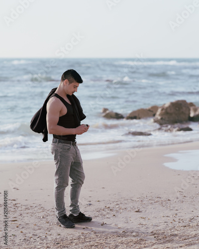 A young athletic man with short dark hair, a dark beard, and a mustache. He is standing in profile on a sandy beach.
